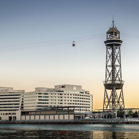 ยูโรสตาร์ แกรนด์ มารีน่า จีเอล โฮเต็ล Hotel บาร์เซโลนา ภายนอก รูปภาพ The photo shows a modern building alongside a waterfront, with a distinctive tower nearby. The tower appears to be part of a cable car system, featuring a conical roof and a series of cables extending from it. The scene is set against a colorful sky,