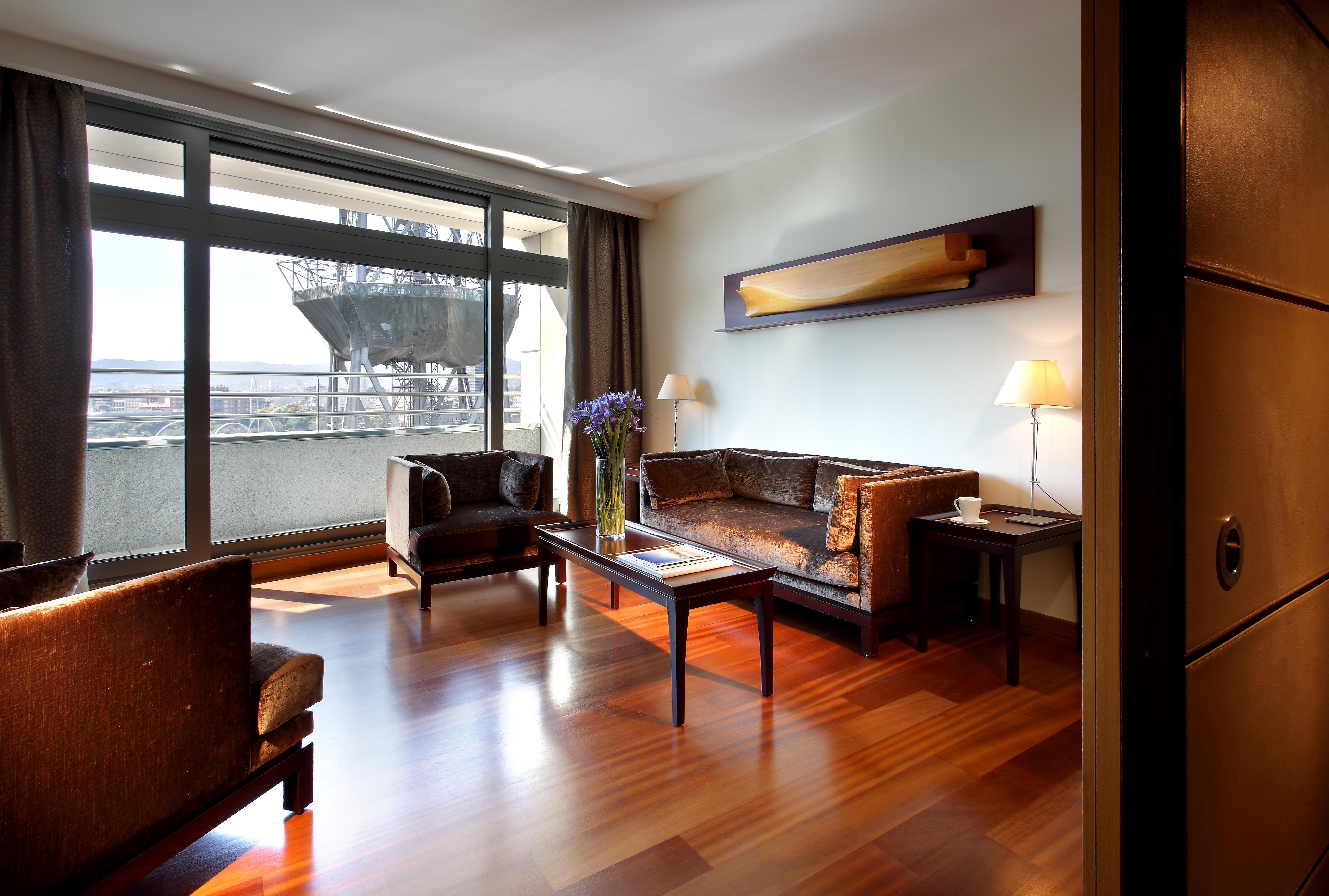 ยูโรสตาร์ แกรนด์ มารีน่า จีเอล โฮเต็ล Hotel บาร์เซโลนา ภายนอก รูปภาพ The photo depicts a modern living room setting. It features a spacious area with large windows allowing natural light to flood in. The room is furnished with two dark chairs and a brown couch, all arranged around a wooden coffee table. A decorative l