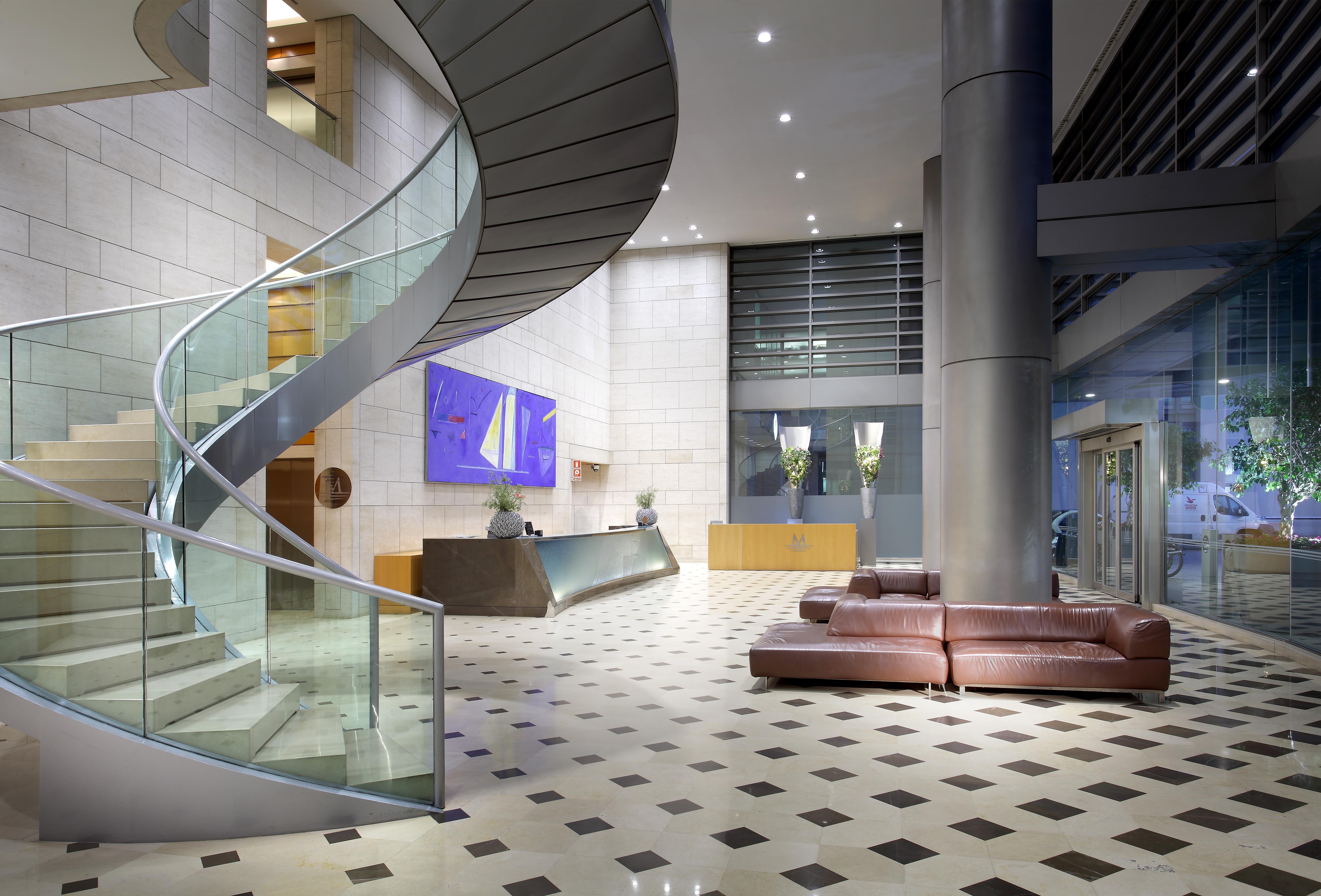 ยูโรสตาร์ แกรนด์ มารีน่า จีเอล โฮเต็ล Hotel บาร์เซโลนา ภายนอก รูปภาพ The photo shows a modern hotel or office lobby featuring a sleek, spiral staircase made of glass and metal. The staircase is accompanied by a large, open space with a tiled floor that has a checkered pattern, combining light and dark tiles. 

To the 