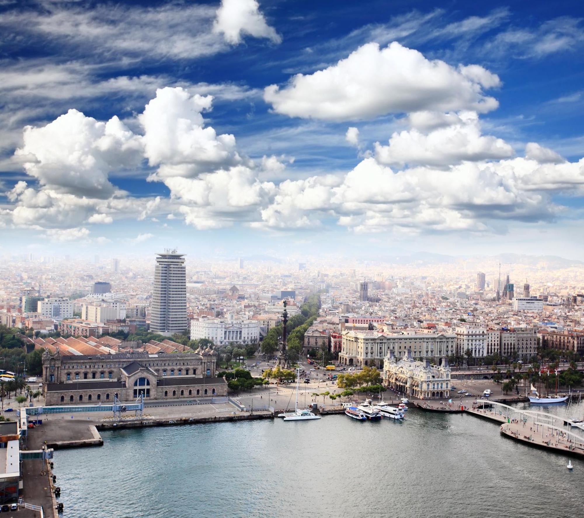 ยูโรสตาร์ แกรนด์ มารีน่า จีเอล โฮเต็ล Hotel บาร์เซโลนา ภายนอก รูปภาพ The image shows a panoramic view of a cityscape, likely featuring a coastal area with a harbor. In the foreground, there are buildings near the water, including a large structure that might be a historic or notable building. The scene extends into th
