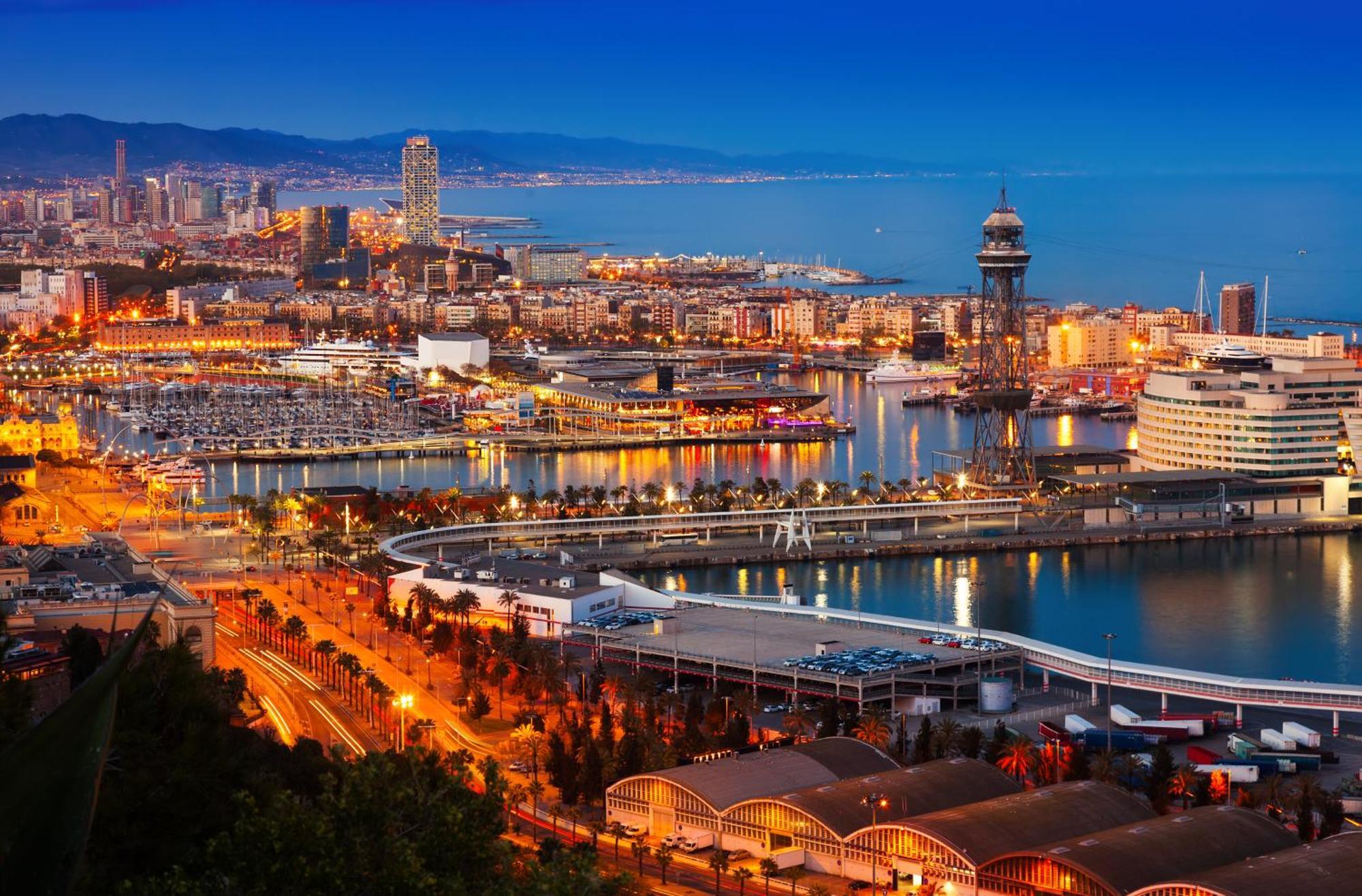 ยูโรสตาร์ แกรนด์ มารีน่า จีเอล โฮเต็ล Hotel บาร์เซโลนา ภายนอก รูปภาพ The photo depicts a scenic view of a coastal city at dusk. In the foreground, there are illuminated buildings and a marina filled with boats. The waterfront features a curved walkway and a dock area. In the background, you can see a skyline with tall