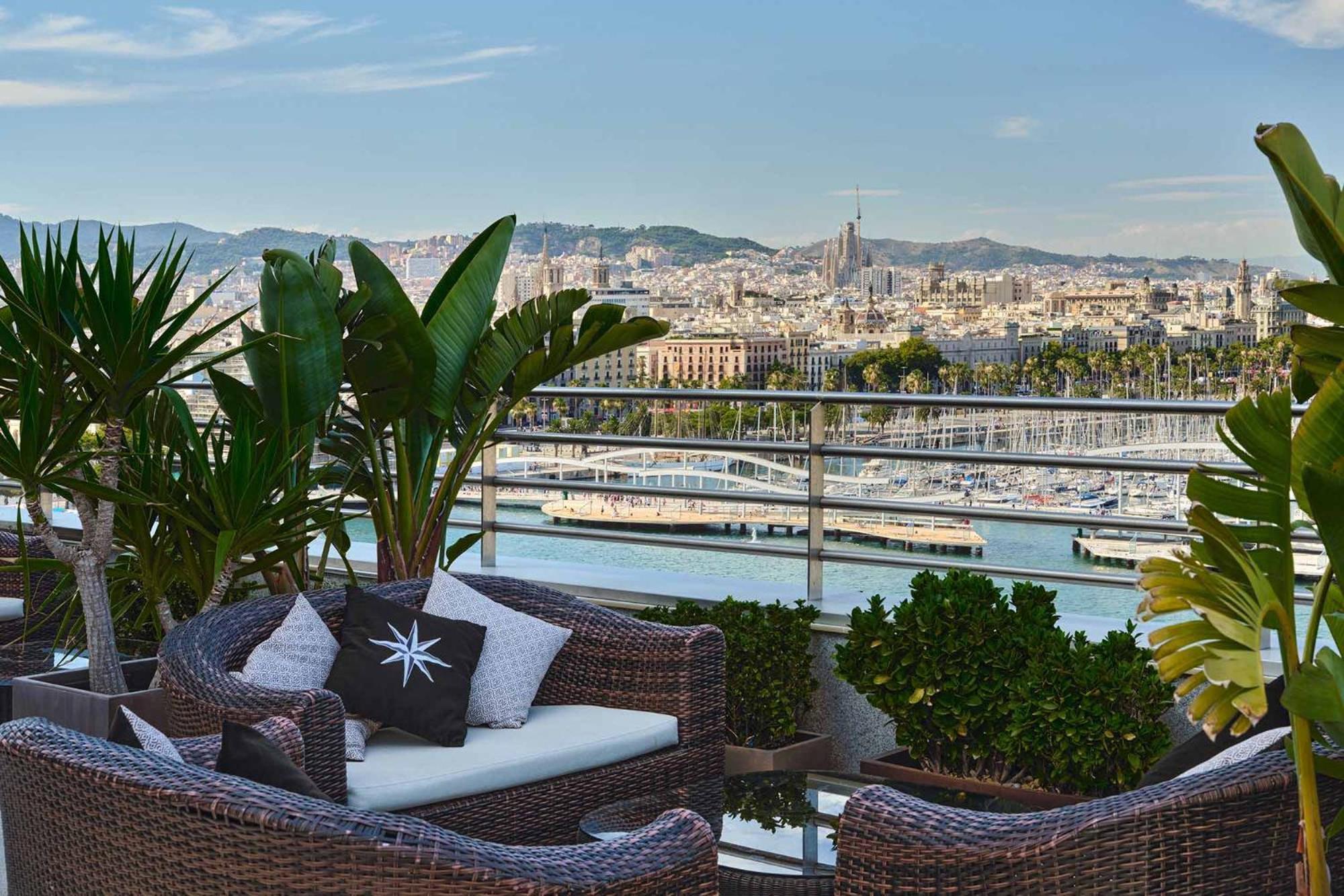 ยูโรสตาร์ แกรนด์ มารีน่า จีเอล โฮเต็ล Hotel บาร์เซโลนา ภายนอก รูปภาพ The photo shows a beautiful outdoor terrace with comfortable, woven furniture, including a sofa and cushions. In the background, there’s a scenic view of a city skyline, likely featuring a harbor or waterfront area, with boats in the water. There are