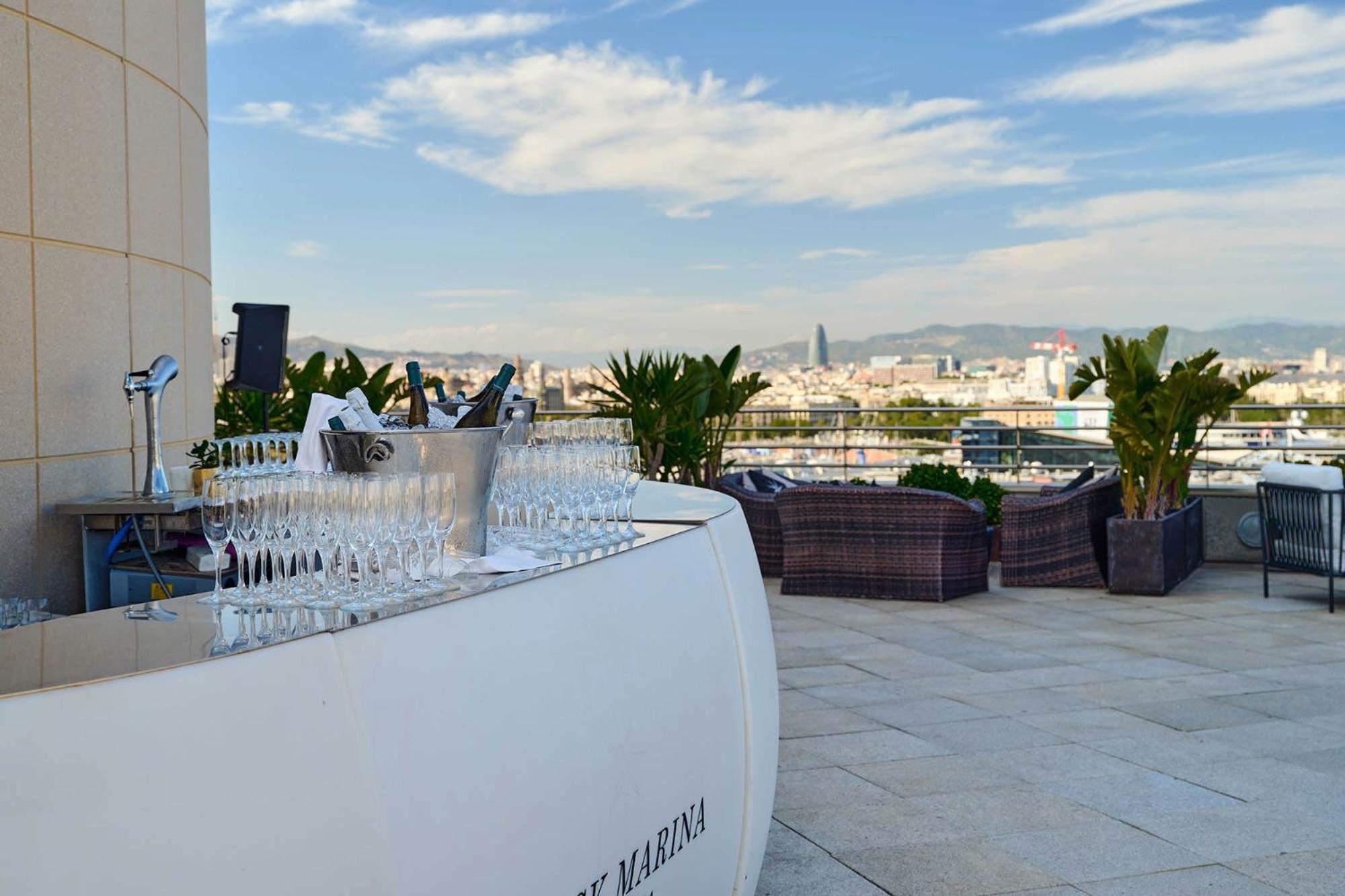 ยูโรสตาร์ แกรนด์ มารีน่า จีเอล โฮเต็ล Hotel บาร์เซโลนา ภายนอก รูปภาพ The photo shows a stylish rooftop bar or terrace area with a modern, curved white bar counter. In front of the bar, there is a row of sparkling glasses lined up for drinks. The background features a beautiful skyline and distant mountains, with a cle