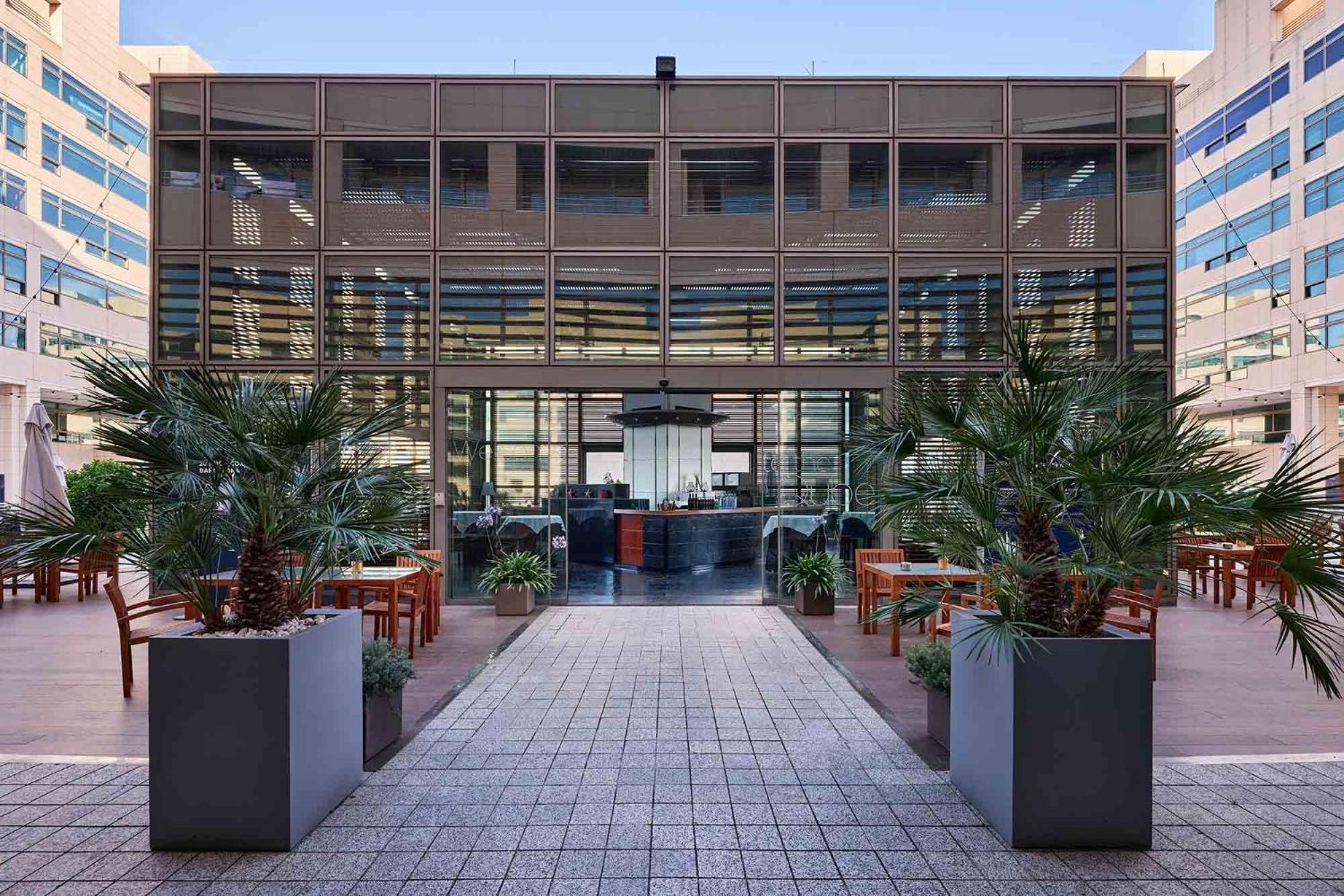ยูโรสตาร์ แกรนด์ มารีน่า จีเอล โฮเต็ล Hotel บาร์เซโลนา ภายนอก รูปภาพ The photo depicts a modern building with a glass facade, featuring a central entrance framed by large planters with tropical plants. The entrance leads to an interior space that appears to be a restaurant or café, with a stylish counter and seating v
