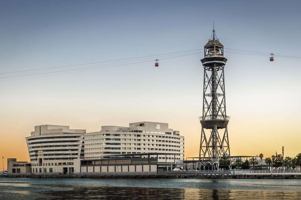 ยูโรสตาร์ แกรนด์ มารีน่า จีเอล โฮเต็ล Hotel บาร์เซโลนา ภายนอก รูปภาพ The photo shows a modern building alongside a waterfront, with a distinctive tower nearby. The tower appears to be part of a cable car system, featuring a conical roof and a series of cables extending from it. The scene is set against a colorful sky,