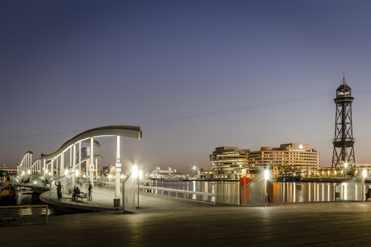 ยูโรสตาร์ แกรนด์ มารีน่า จีเอล โฮเต็ล Hotel บาร์เซโลนา ภายนอก รูปภาพ The photo depicts a scenic waterfront view during twilight. On the left, there is a modern, curved pedestrian walkway that appears to be well-lit, inviting pedestrians. To the right, a tall structure resembling a lighthouse or observation tower stand